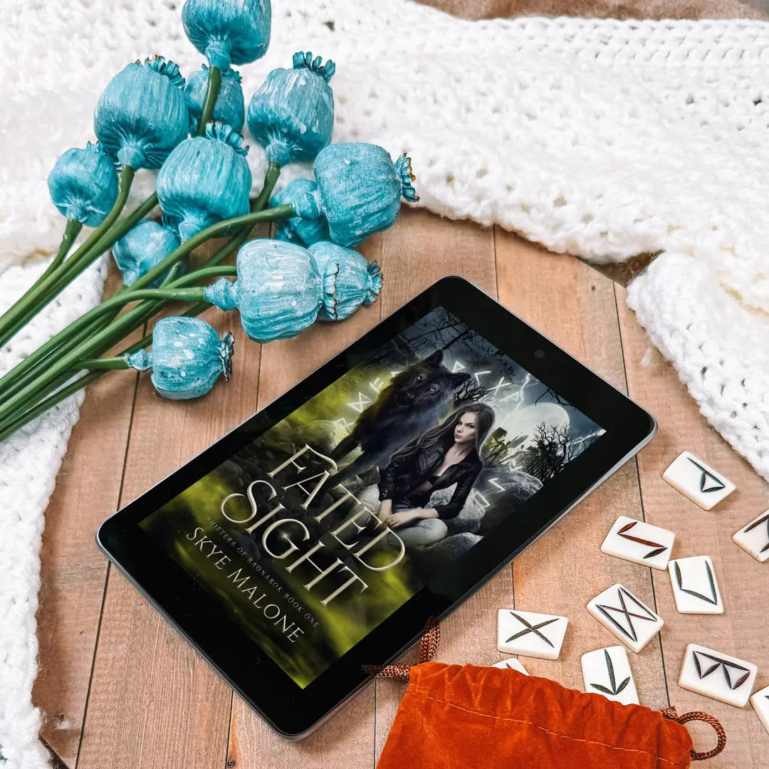 Ebook edition of Fated Sight by Skye Malone lying on a wooden table with blue flowers on the left and white runes falling from a brown bag on the right. A white sweater surrounds the objects.